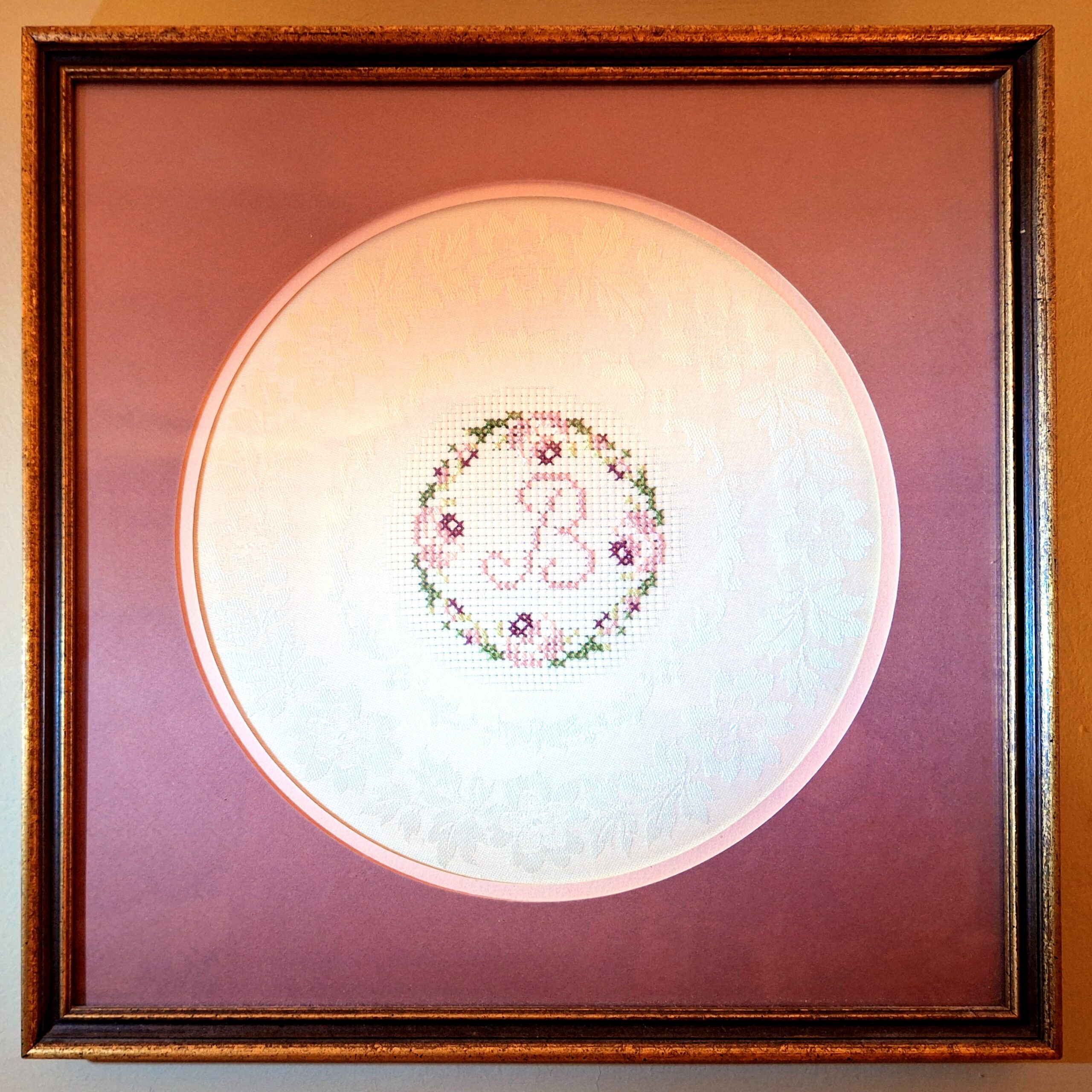 A cross-stitch embroidered capital letter B on white satin, surrounded by a mauve circular-cut matte in a square gold-tone wood frame