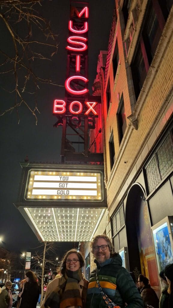 Two people standing outside the marquee of the Music Box Theater at night, with the vertical neon sign above spelling "Music Box"